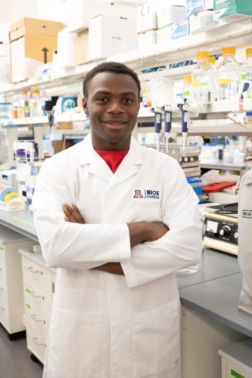 A person in a white lab coat with standing with arms crossed in a laboratory filled with scientific equipment.