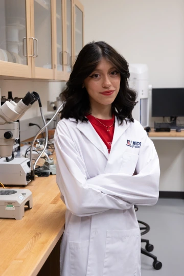A person in a white lab coat with standing with arms crossed in a laboratory filled with scientific equipment.