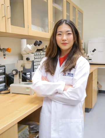 A person in a white lab coat with standing with arms crossed in a laboratory filled with scientific equipment.