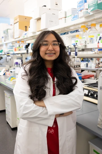 A person in a white lab coat with standing with arms crossed in a laboratory filled with scientific equipment.