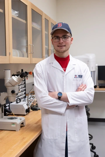 A person in a white lab coat with standing with arms crossed in a laboratory filled with scientific equipment.
