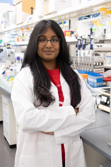 A person in a white lab coat with standing with arms crossed in a laboratory filled with scientific equipment.