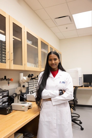A person in a white lab coat with standing with arms crossed in a laboratory filled with scientific equipment.