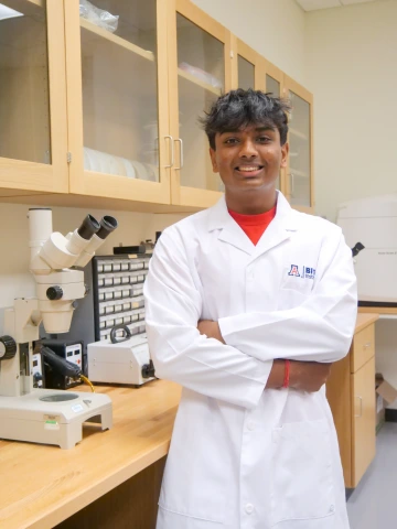 A person in a white lab coat with standing with arms crossed in a laboratory filled with scientific equipment.