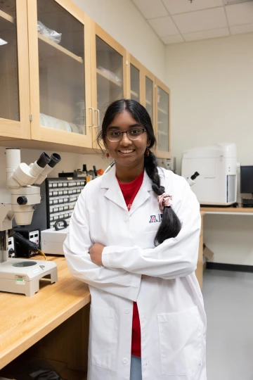 A person in a white lab coat with standing with arms crossed in a laboratory filled with scientific equipment.