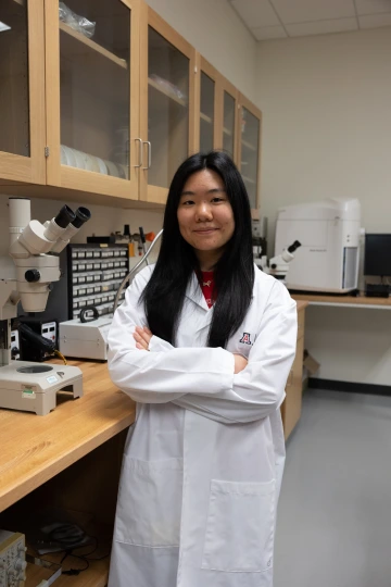 A person in a white lab coat with standing with arms crossed in a laboratory filled with scientific equipment.
