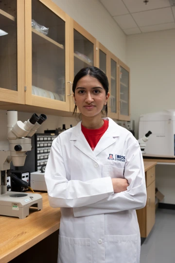 A person in a white lab coat with standing with arms crossed in a laboratory filled with scientific equipment.