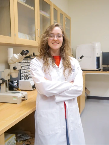 A person in a white lab coat with standing with arms crossed in a laboratory filled with scientific equipment.
