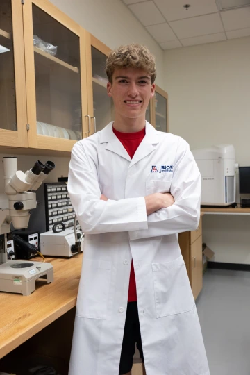 A person in a white lab coat with standing with arms crossed in a laboratory filled with scientific equipment.