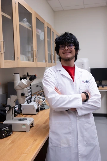 A person in a white lab coat with standing with arms crossed in a laboratory filled with scientific equipment.