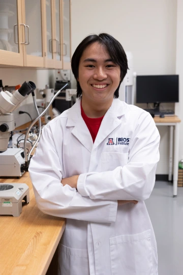 A person in a white lab coat with standing with arms crossed in a laboratory filled with scientific equipment.