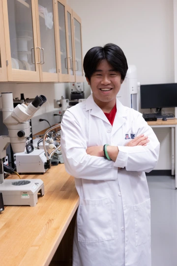 A person in a white lab coat with standing with arms crossed in a laboratory filled with scientific equipment.