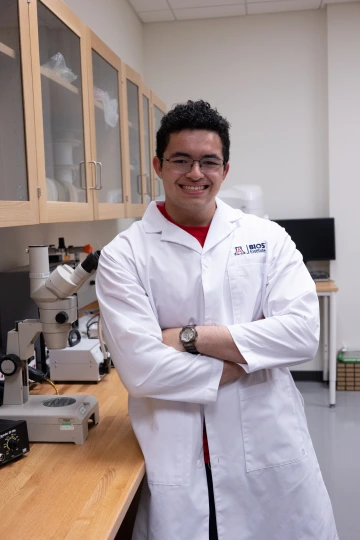 A person in a white lab coat with standing with arms crossed in a laboratory filled with scientific equipment.