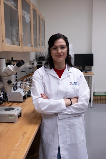 A person in a white lab coat with standing with arms crossed in a laboratory filled with scientific equipment.