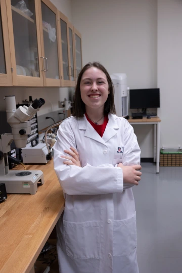 A person in a white lab coat with standing with arms crossed in a laboratory filled with scientific equipment.