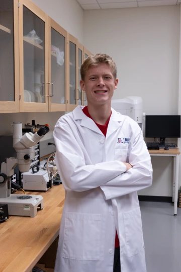 A person in a white lab coat with standing with arms crossed in a laboratory filled with scientific equipment.
