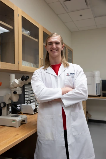 A person in a white lab coat with standing with arms crossed in a laboratory filled with scientific equipment.