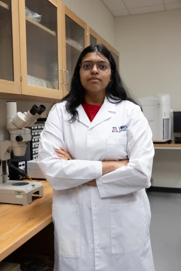A person in a white lab coat with standing with arms crossed in a laboratory filled with scientific equipment.