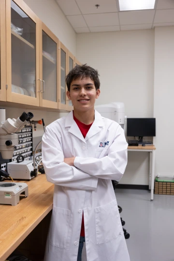 A person in a white lab coat with standing with arms crossed in a laboratory filled with scientific equipment.