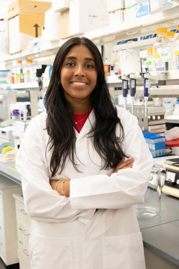 A person in a white lab coat with standing with arms crossed in a laboratory filled with scientific equipment.