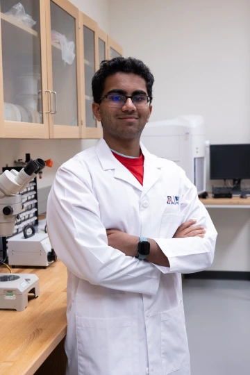 A person in a white lab coat with standing with arms crossed in a laboratory filled with scientific equipment.