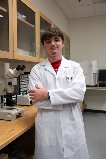 A person in a white lab coat with standing with arms crossed in a laboratory filled with scientific equipment.