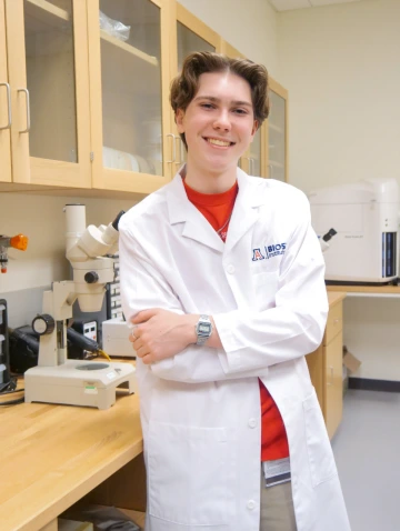 A person in a white lab coat with standing with arms crossed in a laboratory filled with scientific equipment.