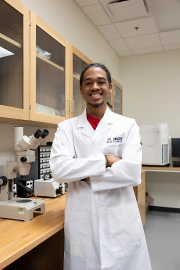 A person in a white lab coat with standing with arms crossed in a laboratory filled with scientific equipment.