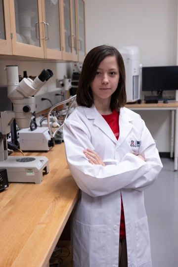 A person in a white lab coat with standing with arms crossed in a laboratory filled with scientific equipment.