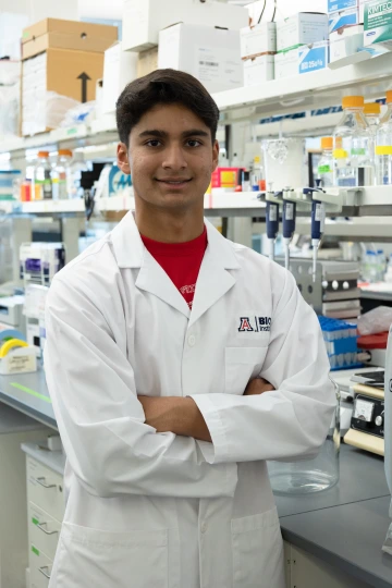 A person in a white lab coat with standing with arms crossed in a laboratory filled with scientific equipment.