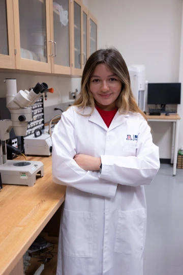 A person in a white lab coat with standing with arms crossed in a laboratory filled with scientific equipment.