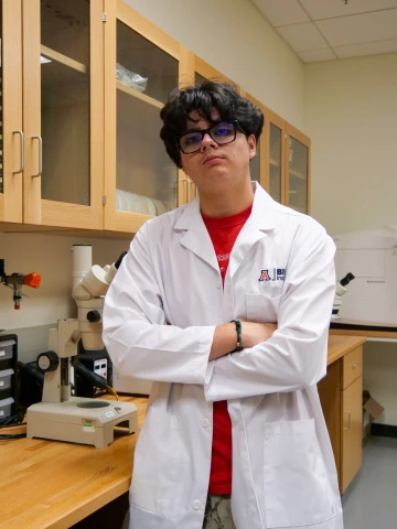 A person in a white lab coat with standing with arms crossed in a laboratory filled with scientific equipment.