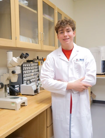 A person in a white lab coat with standing with arms crossed in a laboratory filled with scientific equipment.