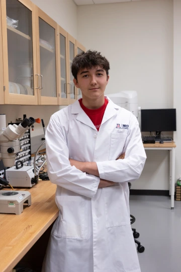 A person in a white lab coat with standing with arms crossed in a laboratory filled with scientific equipment.