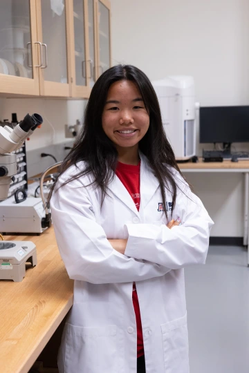A person in a white lab coat with standing with arms crossed in a laboratory filled with scientific equipment.