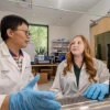 Two people discuss in a lab wearing white coats and blue latex gloves