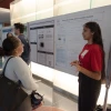 Two individuals engage in a discussion beside a research poster. One person, standing, gestures towards the poster, while the other listens attentively. Several other attendees can be seen in the background.